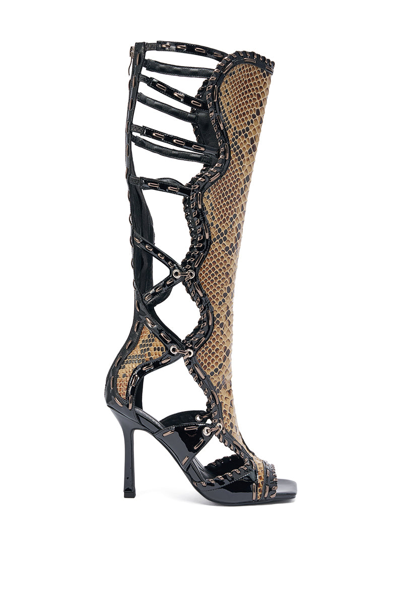 The AKIRA GOLD INDRANI BLACK HIGH SHAFT GLADIATOR SANDAL is a knee-high, open-toe stiletto boot with black cutout straps, a faux patent leather finish, and intricate gladiator-inspired cage detailing. 8