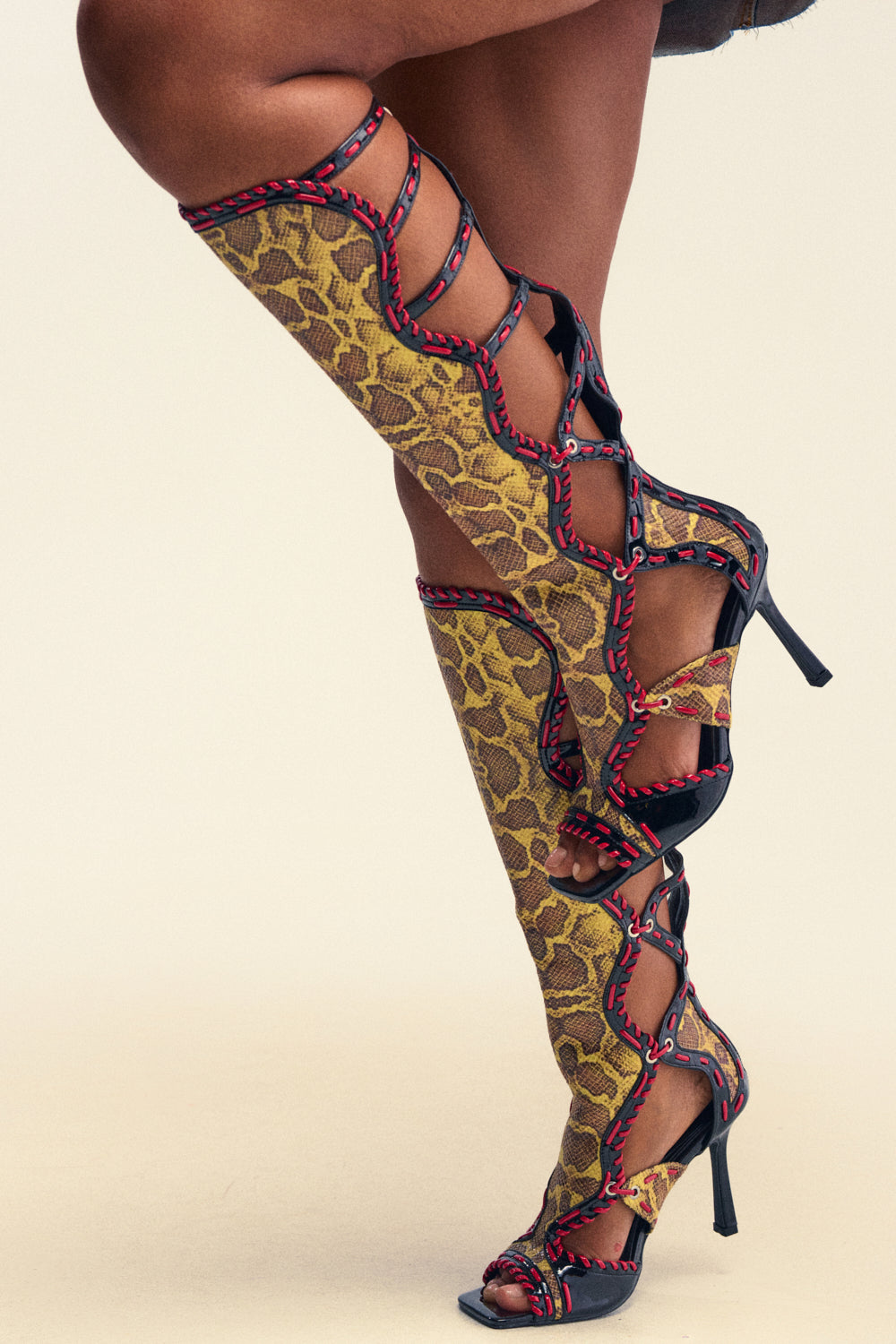 A person wearing the AZALEA WANG INDRANI HIGH SHAFT YELLOW SNAKE PRINT GLADIATOR SANDAL, featuring knee-high open-toe stilettos with a vibrant yellow snakeskin print, black straps, and bold red stitching. Only their legs are visible in the image.
