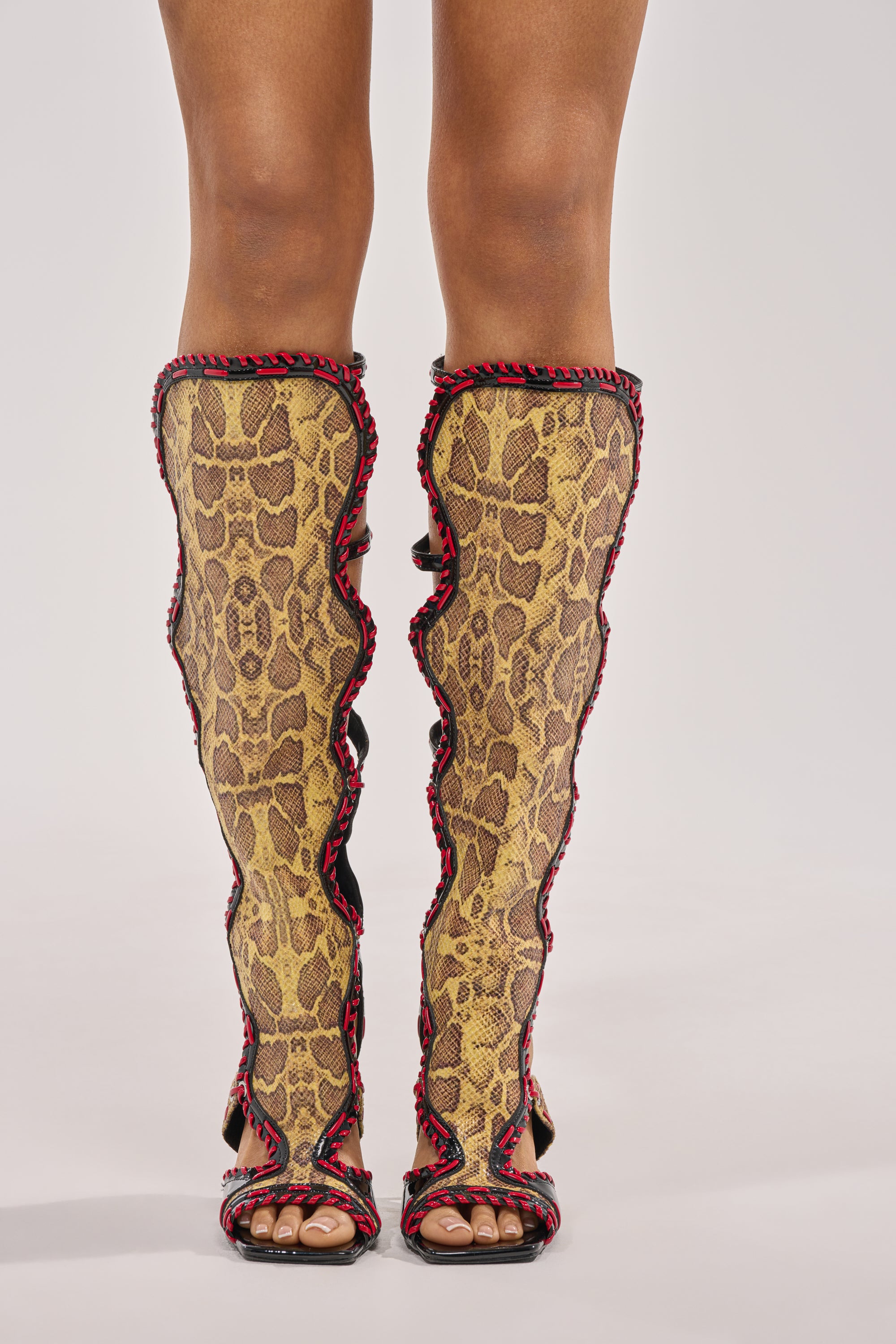 A person wears the AZALEA WANG INDRANI HIGH SHAFT YELLOW SNAKE PRINT GLADIATOR SANDAL with open toes, knee-high yellow and brown snakeskin pattern, black and red trim, and stiletto heels, standing against a plain white background. 1