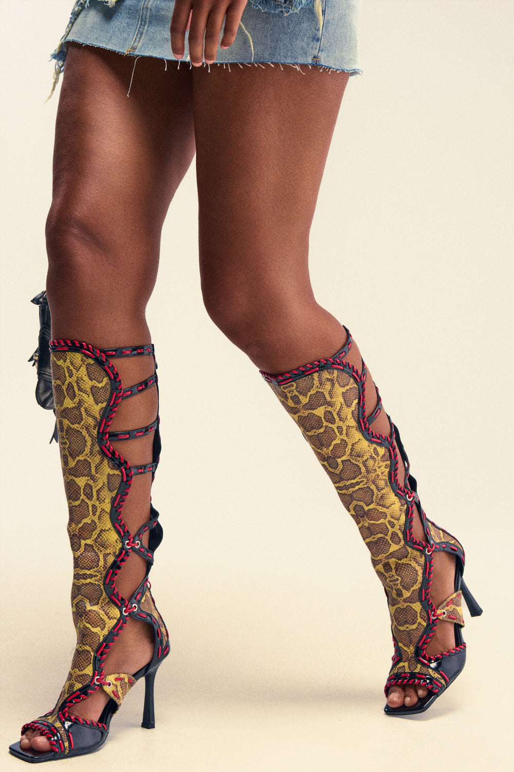 A person wearing a frayed denim mini skirt and the AZALEA WANG INDRANI HIGH SHAFT YELLOW SNAKE PRINT GLADIATOR SANDAL, featuring yellow snakeskin print with red accents, is standing with legs crossed.