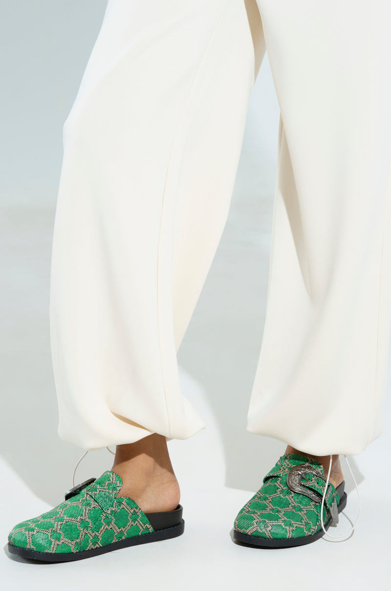 A stylish individual wears crisp white pants with AZALEA WANG INFORMER GREEN SNAKE CLOGS featuring a vibrant snakeskin pattern and large buckles, set against a minimal white backdrop. The clogs include a cushioned insole for extra comfort.