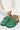 A person wearing cream-colored pants sports AZALEA WANG INFORMER GREEN SNAKE CLOGs with chic snakeskin pattern, large buckles, a black sole, and cushioned insoles, complemented by white ties elegantly wrapped around the pants.