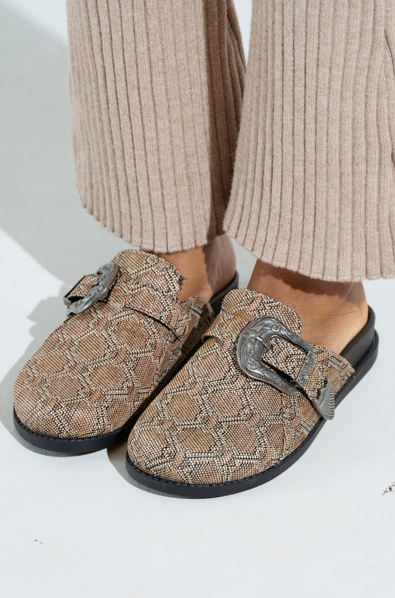 A person wears tan knitted pants and AZALEA WANG INFORMER TAN SNAKE CLOG slip-ons, featuring a striking snakeskin pattern with large decorative buckles, black soles, styled elegantly in natural light on a plain surface.