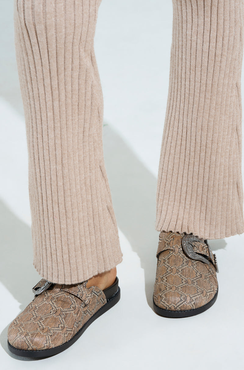 A person wears beige ribbed flare pants and AZALEA WANG INFORMER TAN SNAKE CLOGS with a striking brown and black snakeskin pattern, featuring a strap across the top, on a light background.