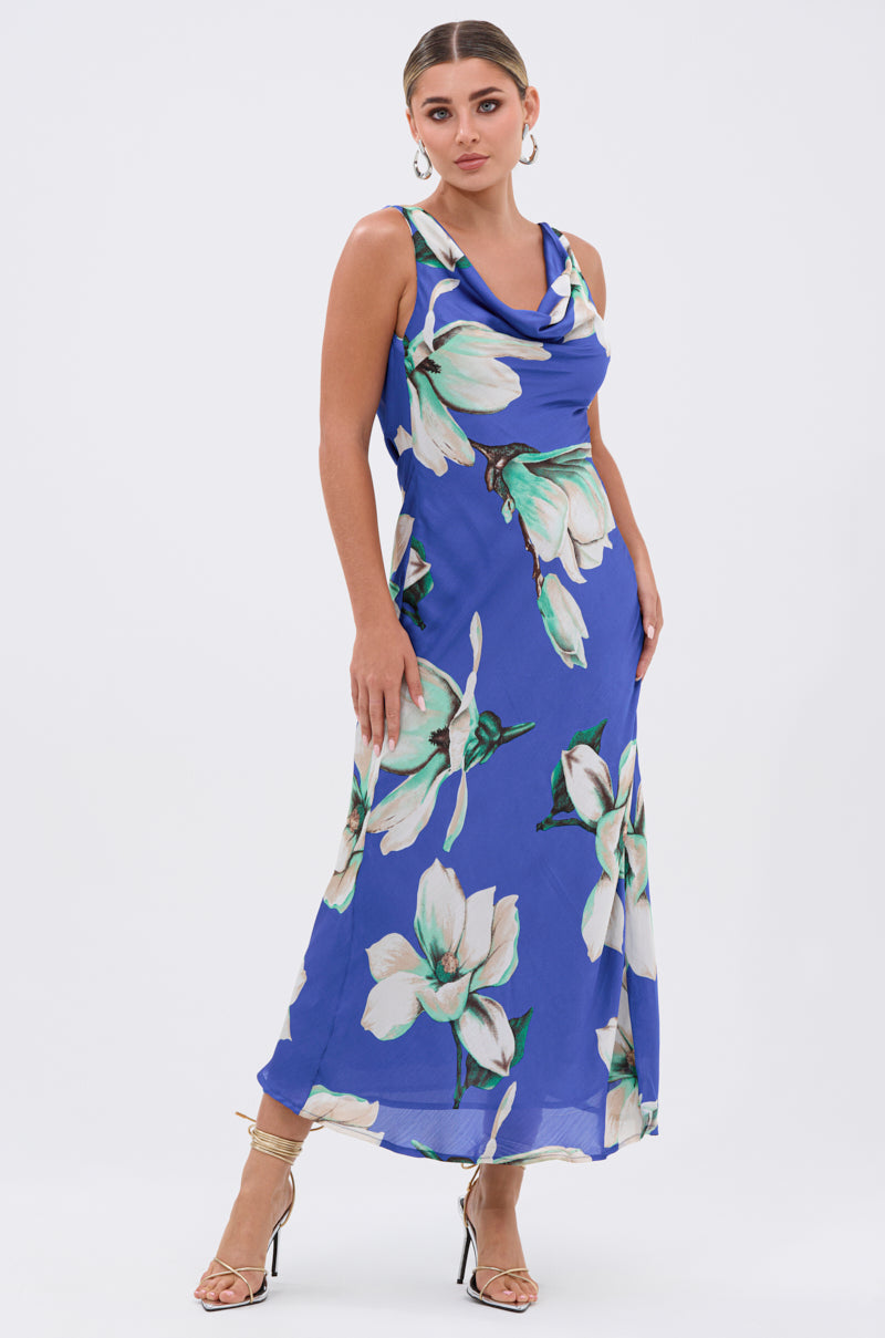 Wearing the SOMETHING SWEET FLORAL MAXI DRESS, a woman with slicked-back hair, hoop earrings, and strappy heels stands confidently, creating an effortless summer wedding look in this sleeveless ankle-length dress with bold white blooms.