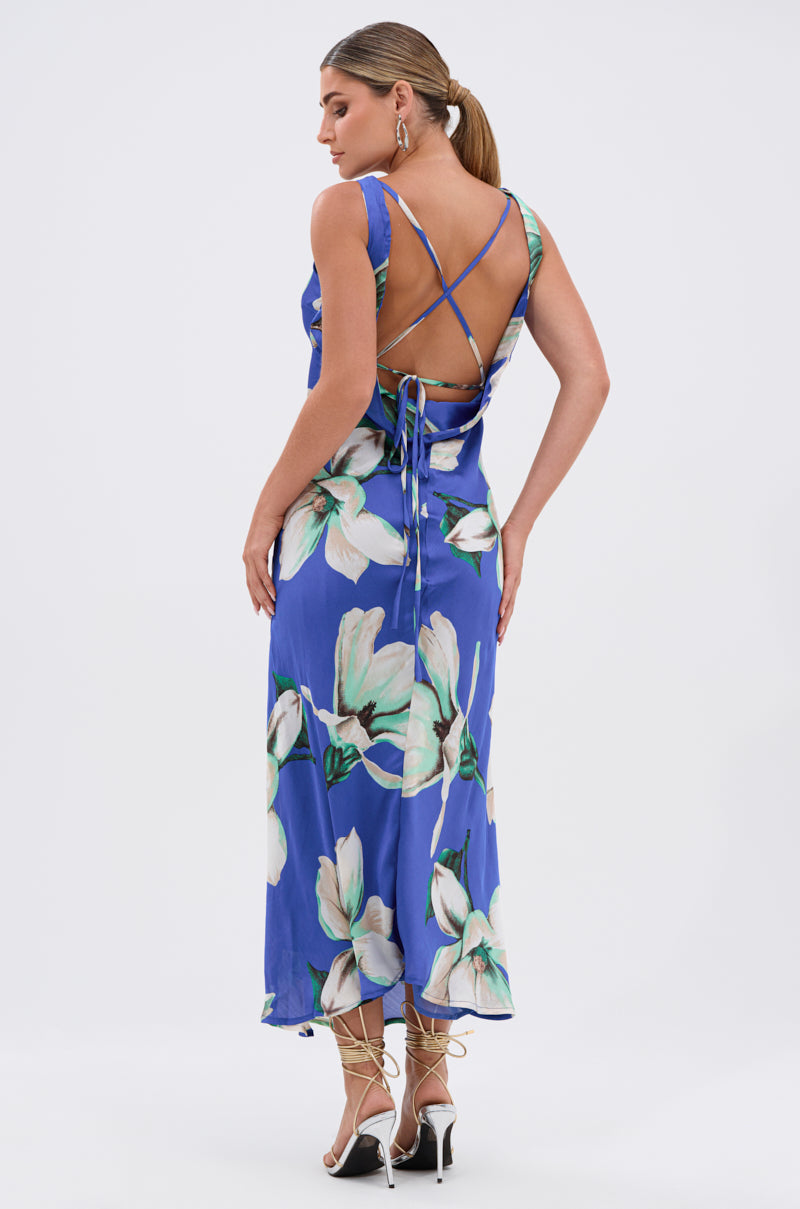 A woman stands facing away from the camera in the SOMETHING SWEET FLORAL MAXI DRESS, a blue sleeveless chiffon dress with bold white floral prints and crisscross straps, paired with strappy high-heeled sandals.