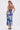 A woman stands facing away from the camera in the SOMETHING SWEET FLORAL MAXI DRESS, a blue sleeveless chiffon dress with bold white floral prints and crisscross straps, paired with strappy high-heeled sandals.