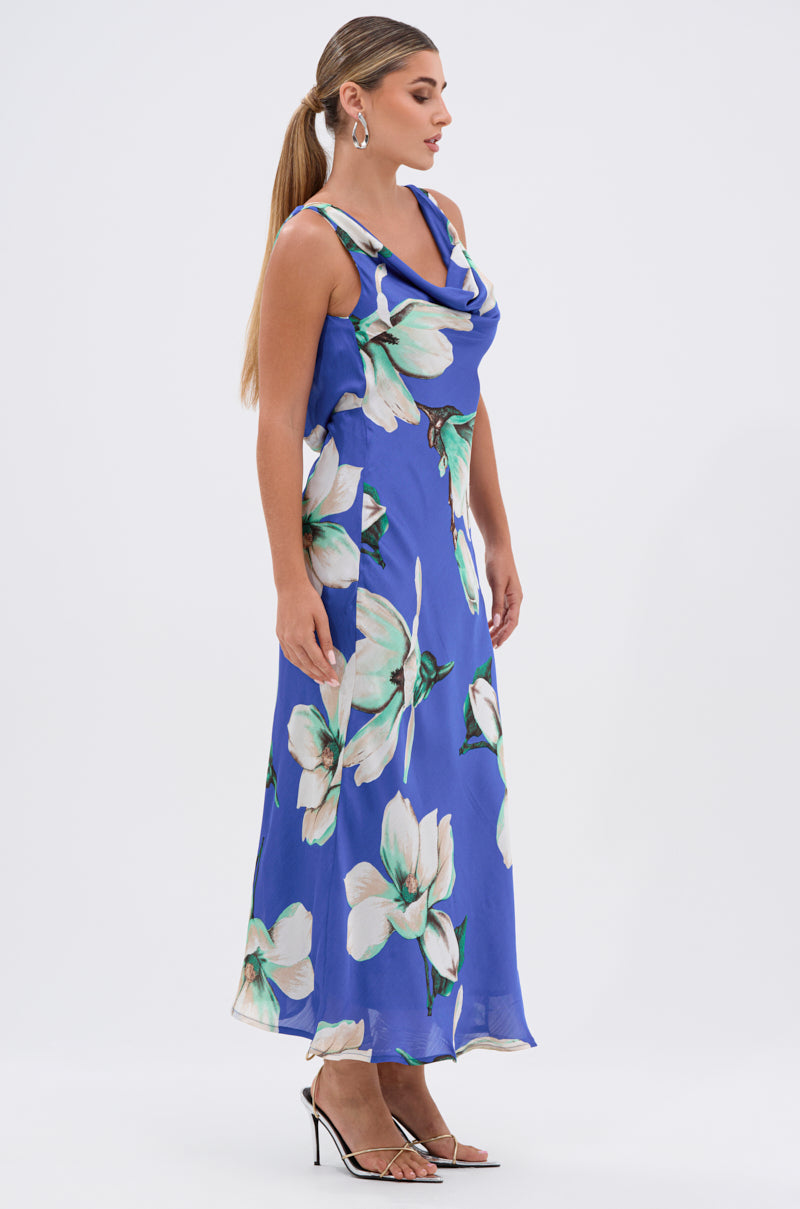 A woman in profile wears the SOMETHING SWEET FLORAL MAXI DRESS, a sleeveless, ankle-length dress with a bold white floral print, paired with hoop earrings and strappy high-heeled sandals. Her long hair is styled in a sleek ponytail.