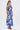 A woman in profile wears the SOMETHING SWEET FLORAL MAXI DRESS, a sleeveless, ankle-length dress with a bold white floral print, paired with hoop earrings and strappy high-heeled sandals. Her long hair is styled in a sleek ponytail.