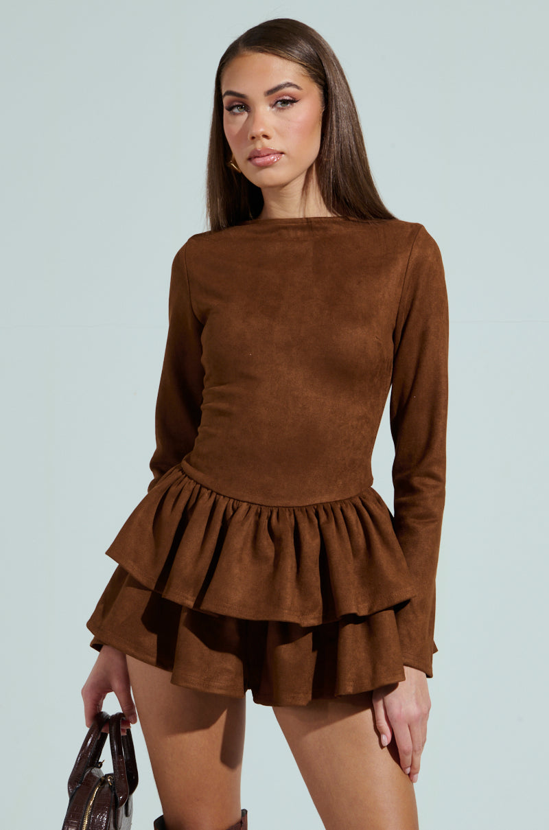 A woman in a brown suede ruffle long-sleeve romper stands against a plain background, her long hair cascading down as she holds a textured brown handbag. Knee-high suede boots complete her chic look.