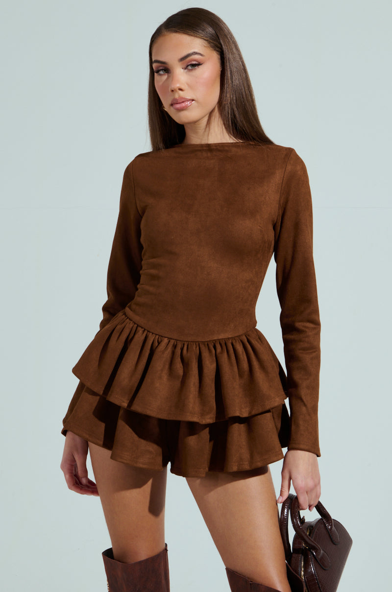 Against a light background, a person exudes confidence in the SUEDE RUFFLE LONG SLEEVE ROMPER with a scoop neckline. They hold a brown handbag and complete the look with knee-high brown boots, radiating poise.