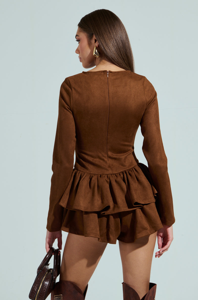 A woman with long brown hair, back to the camera, wears a SUEDE RUFFLE LONG SLEEVE ROMPER featuring a peplum hem. She pairs it with matching brown boots and carries a small handbag against a light blue backdrop.