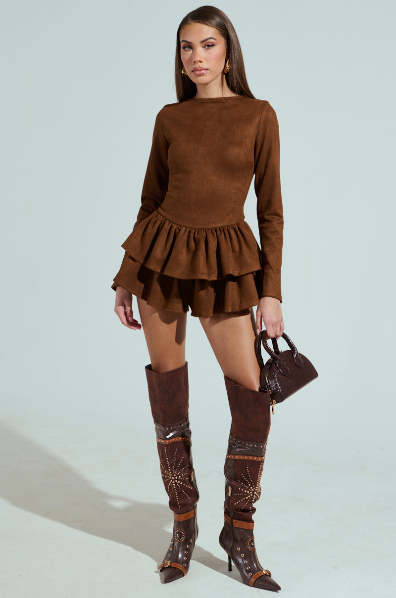A person in a SUEDE RUFFLE LONG SLEEVE ROMPER with a scoop neckline poses against a light background, holding a small black purse and wearing tall dark brown cowboy boots embellished with metallic details.