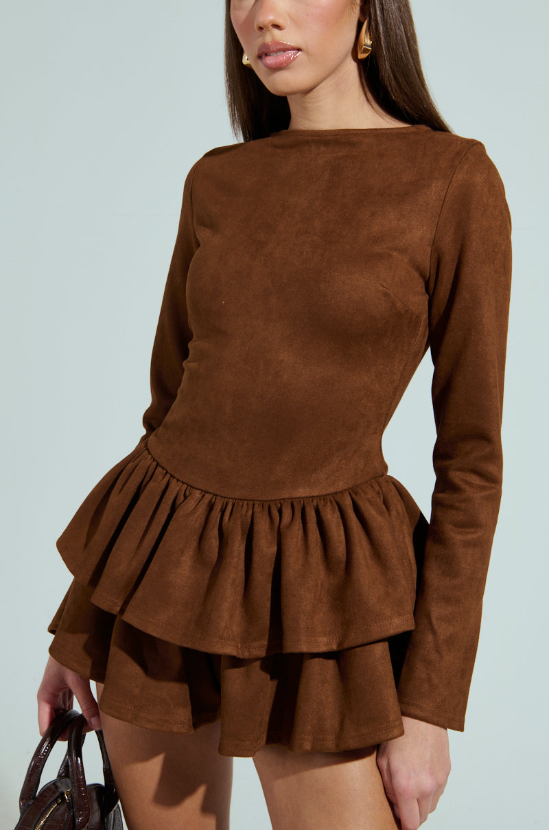 Wearing a SUEDE RUFFLE LONG SLEEVE ROMPER, a woman elegantly holds a small handbag. Her outfit boasts a fitted bodice and ruffled skirt, enhancing her style. Her long hair flows over delicate earrings against a light blue background.