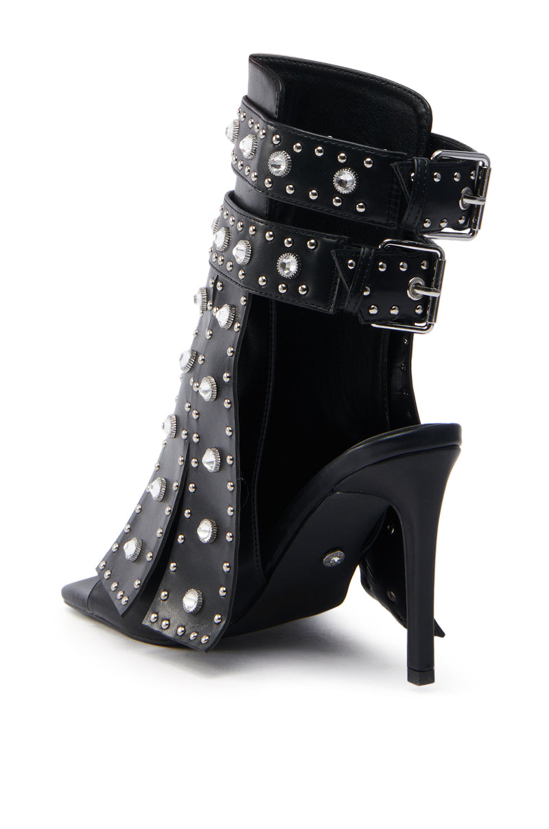 The AZALEA WANG IRELYN BLACK EMBELLISHED SANDAL is a black faux leather high-heeled ankle bootie with open sides, featuring two wide straps with large silver buckles around the ankle and adorned with numerous silver studs, complementing its slender stiletto heel.