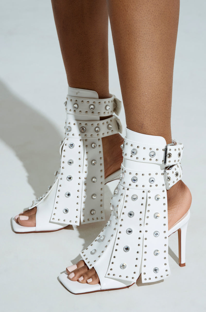 Close-up of a person wearing the AZALEA WANG IRELYN WHITE EMBELLISHED SANDAL, featuring elegant stiletto heels with silver studs and hardware. The faux leather bootie's multiple straps and peep toes beautifully contrast against a light background.