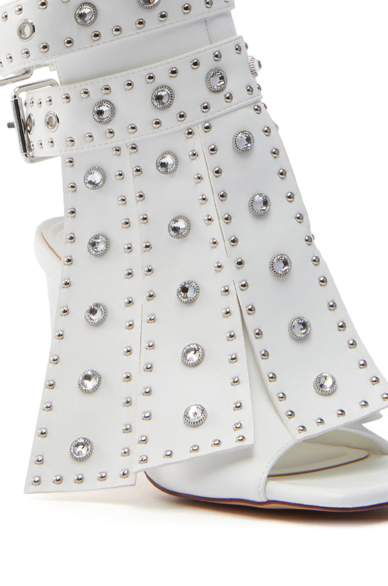 Close-up of an AZALEA WANG IRELYN WHITE EMBELLISHED SANDAL with an open toe and metallic studs, featuring circular decorations and a wide ankle strap with a buckle. The wedge heel adds to its edgy appeal.