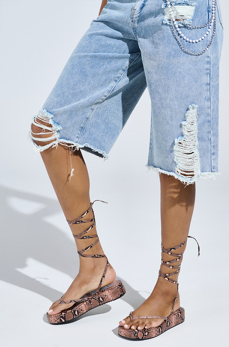 The person sports distressed, ripped denim bermuda shorts and AKIRA Irvine Brown Snake Lace Up Flat Sandals with a chic flatform sole. A silver chain accessory adds flair to the ensemble against a light-colored background.