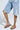 A person wearing distressed denim shorts with rips and chains stands casually against a light background, their legs crossed. They're sporting the AKIRA IRVINE BROWN SNAKE LACE UP FLAT SANDALS.
