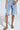 Someone wears light blue ripped denim Bermuda shorts with chain details, paired with AKIRA IRVINE BROWN SNAKE LACE UP FLAT SANDALS. The setting features a plain light gray background.