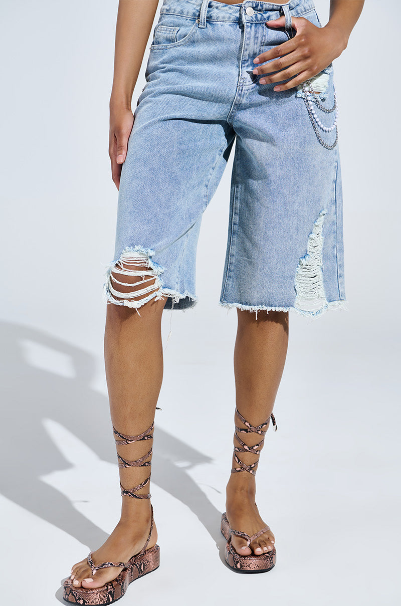 Someone wearing ripped denim Bermuda shorts with chain details, paired with the AKIRA IRVINE BROWN SNAKE LACE UP FLAT SANDAL. The plain background highlights the casual outfit.