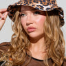 A person with long, wavy hair is wearing the CHEETAH GURLLL BUCKET HAT and posing confidently. They have a thoughtful expression and are dressed in a black top adorned with colorful rhinestone detailing.