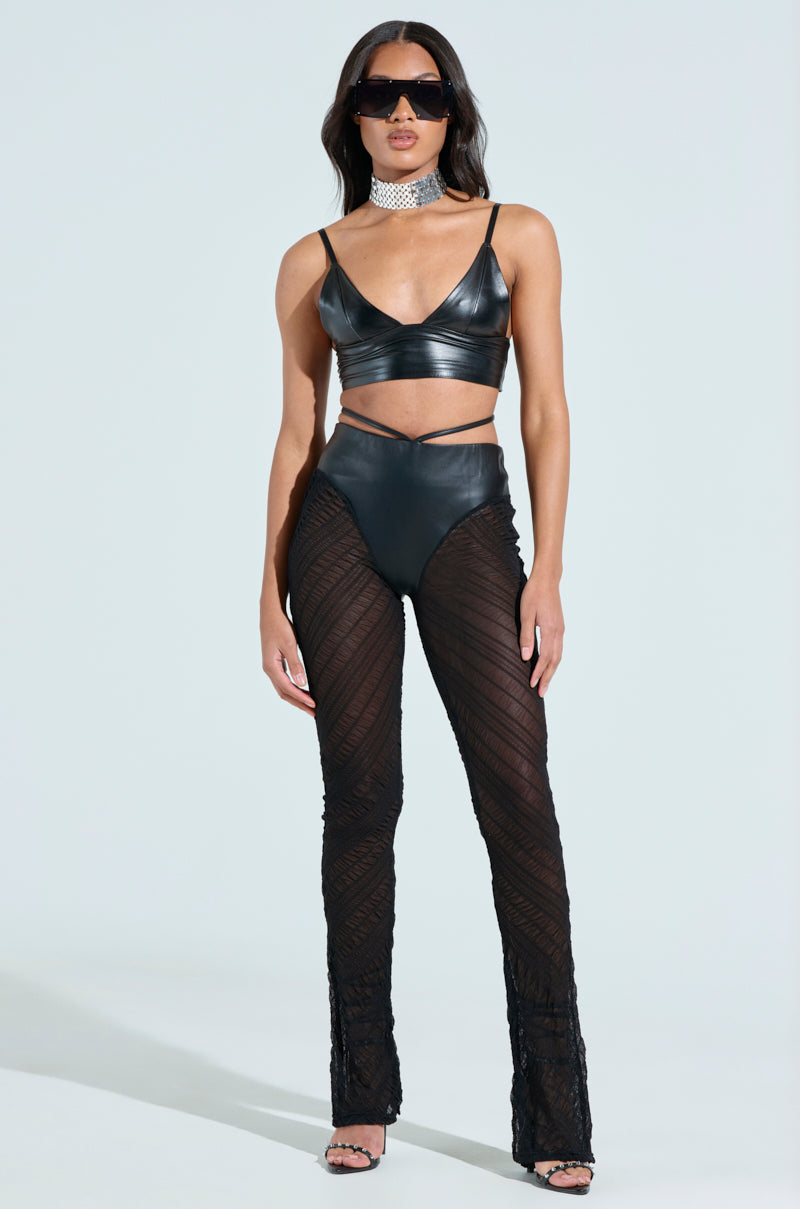 A woman confidently poses in the shiny black IN THE FEELS LACE FLARE PANT and a matching leather crop top. She's accessorized with large sunglasses, a sparkly choker necklace, and strappy high heels. The light neutral background enhances the elegance of her mesh outfit.