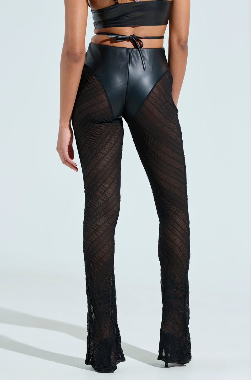 A person in an IN THE FEELS LACE FLARE PANT, featuring a sheer, patterned design revealing skin, stands against a plain, light background. They wear a black leather and mesh sleeveless top that pairs perfectly with the high-rise pants from the same collection.