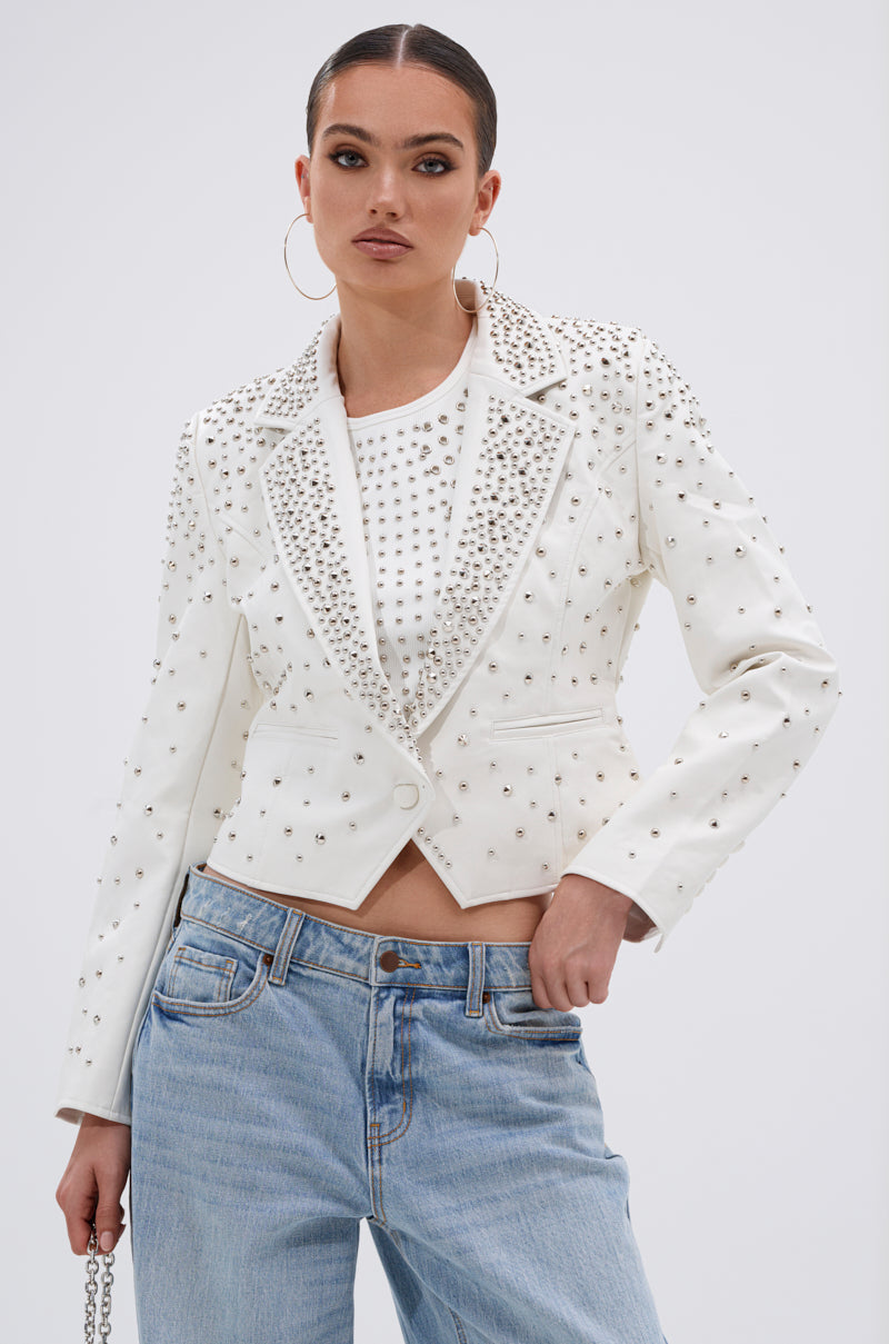 A woman confidently poses against a plain background wearing the VIVA VEGAS FITTED MOTO JACKET over a white top and light blue jeans. She accessorizes with hoop earrings and has slicked-back hair, one hand in her pocket.