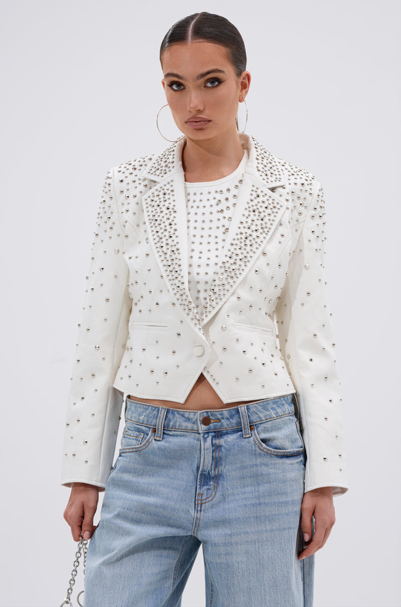 A woman wears the VIVA VEGAS FITTED MOTO JACKET over a white top and blue jeans, standing facing forward with large hoop earrings and a neutral expression against a plain light background.