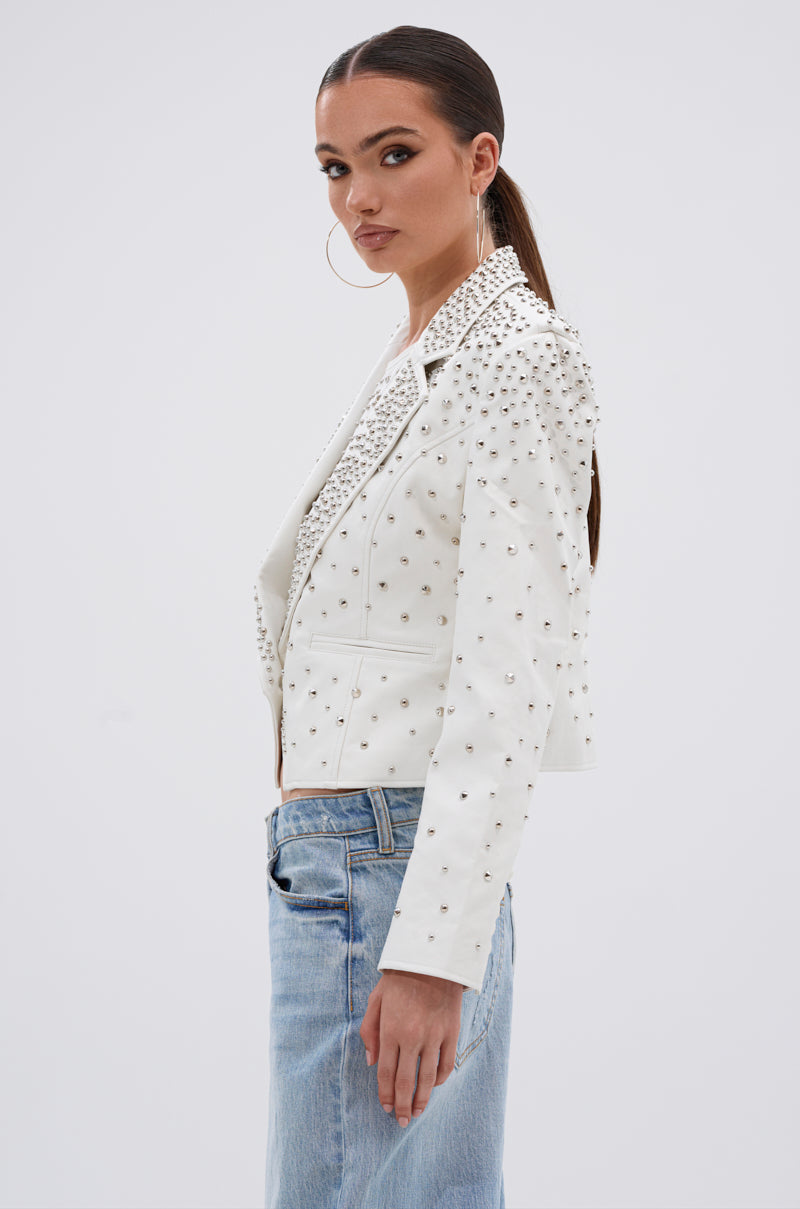 A woman with dark hair in a sleek ponytail models the VIVA VEGAS FITTED MOTO JACKET, featuring stud details, paired with a light blue denim skirt as she poses sideways against a plain light background.
