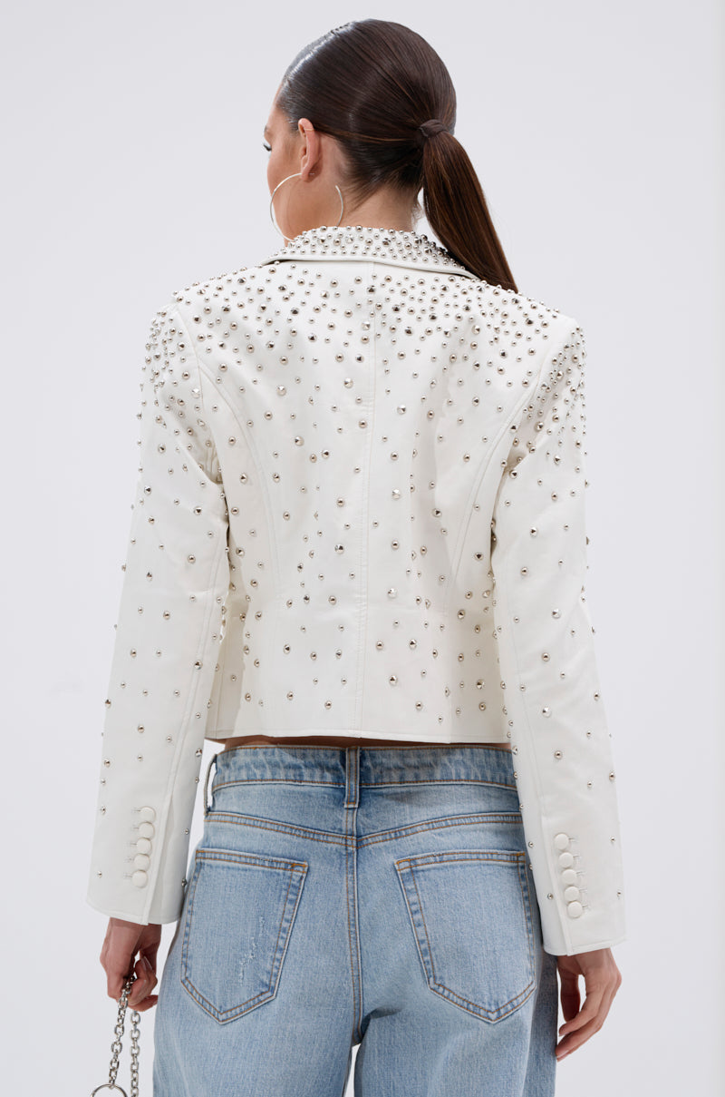 A woman with a sleek ponytail is seen from behind wearing the VIVA VEGAS FITTED MOTO JACKET with silver studs, paired with light blue high-waisted jeans and holding a small chain strap purse in her left hand.