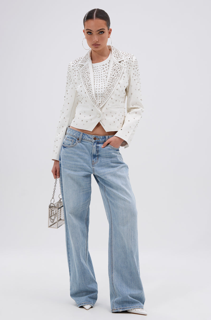 A woman poses against a plain background wearing the VIVA VEGAS FITTED MOTO JACKET over a white top, light blue baggy jeans, white pointed shoes, silver handbag, hoop earrings, and has her hair pulled back.