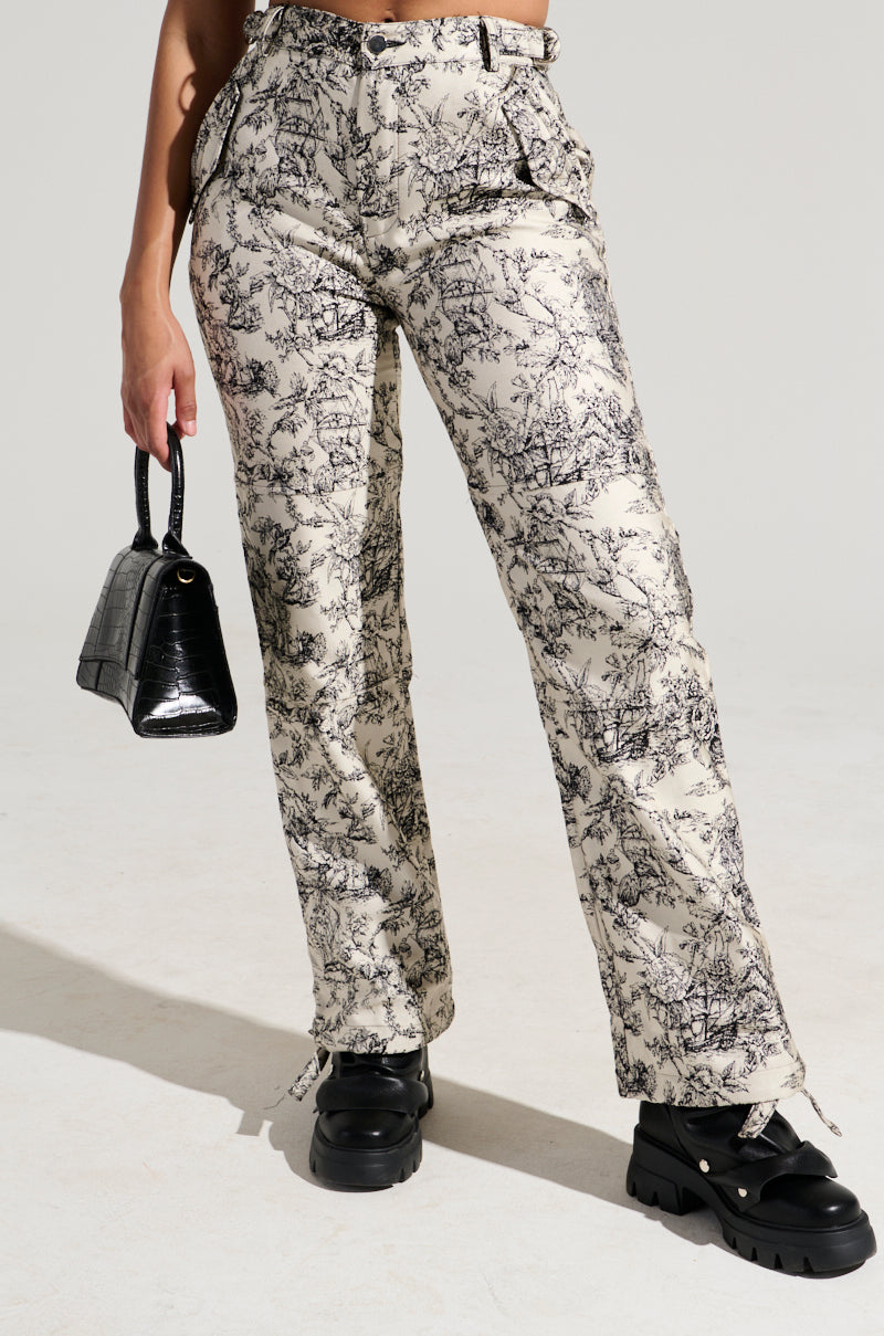 A person stands holding a black handbag, wearing the LET ME GO TROUSER with a wide leg silhouette and high-rise design featuring a black and white nature-inspired print. The ensemble is completed with chunky black platform shoes against a plain light grey background.