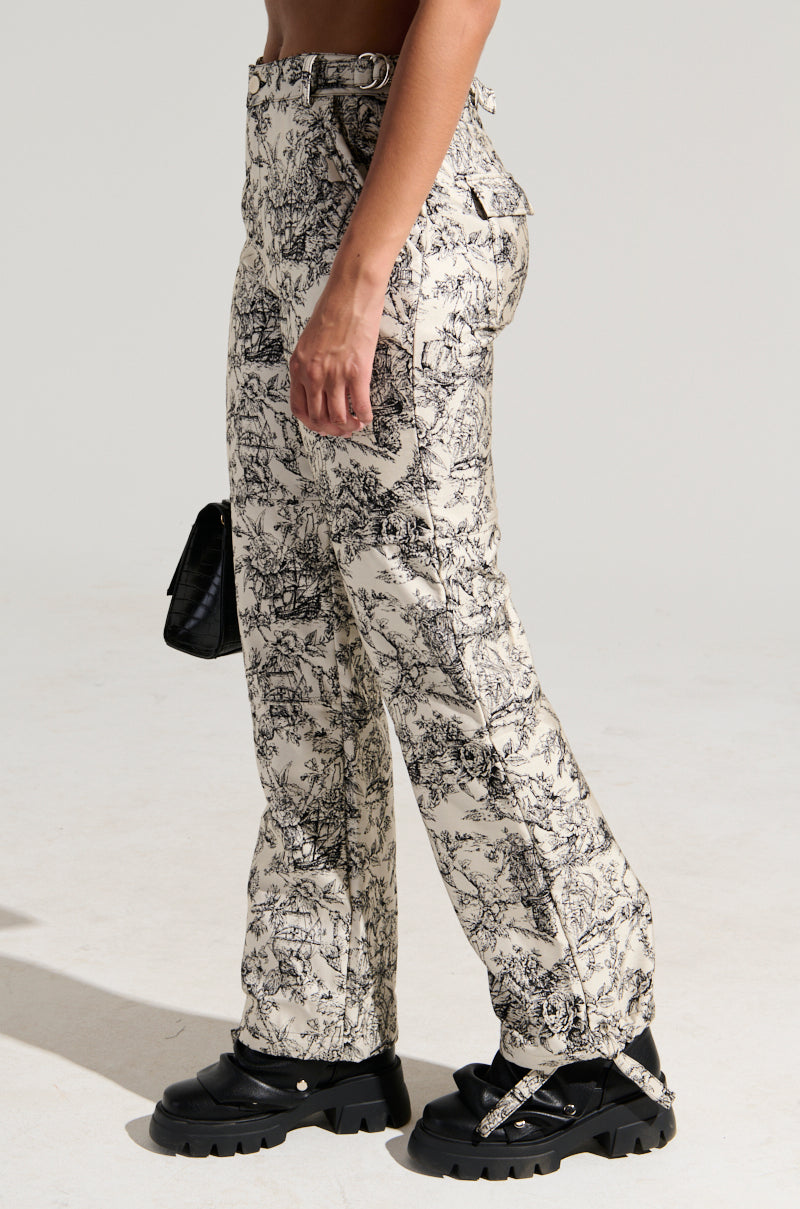 A person is wearing high-waisted, beige LET ME GO TROUSER featuring a black abstract pattern and black chunky sandals. They are holding a small black handbag. The wide leg silhouette of the trousers adds an extra touch of flair. The person's upper body is not visible against the plain, light-colored background.