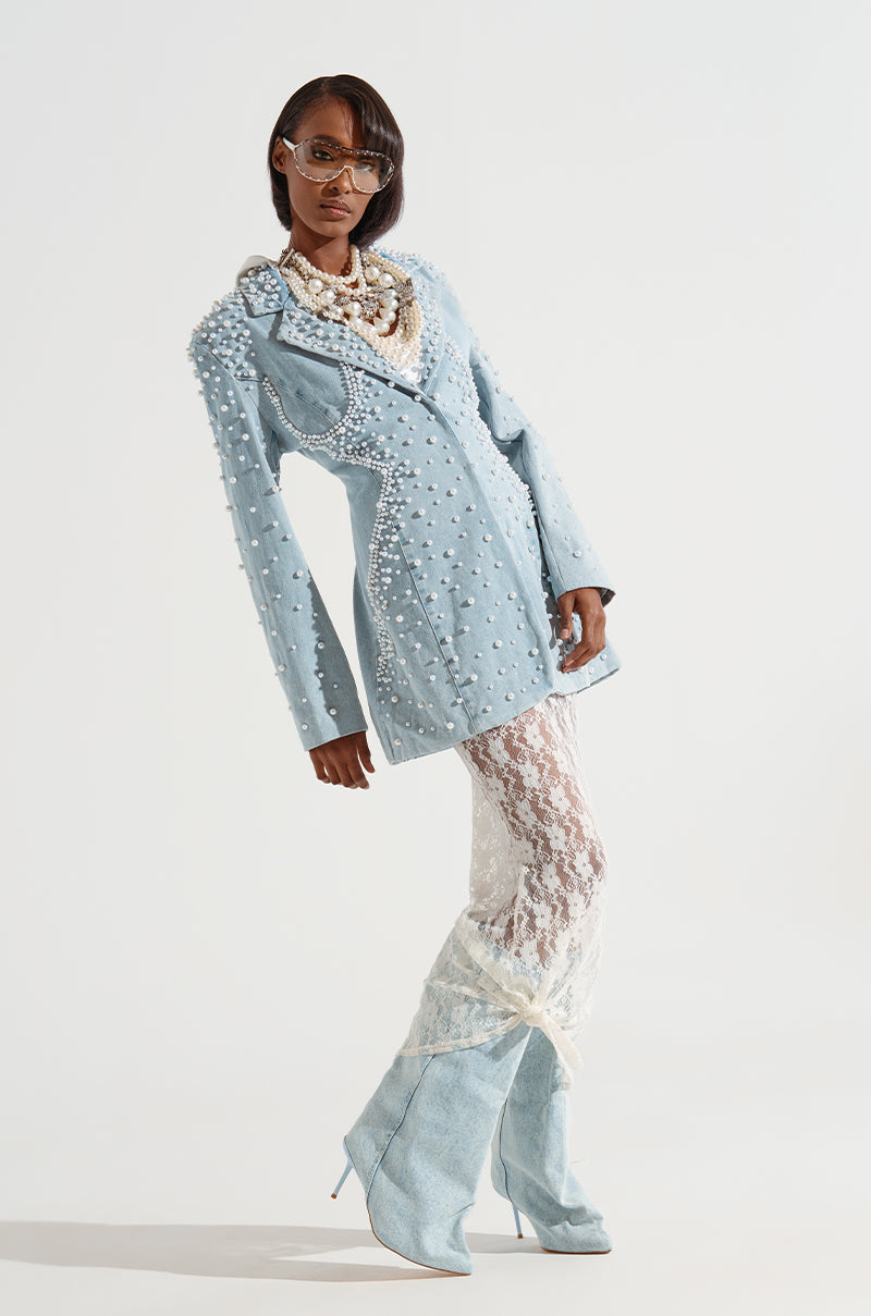 A person poses confidently in an elaborate fashion ensemble featuring the light blue ALIX PEARL EMBELLISHED BLAZER from the AKIRA Label, translucent white lace pants, and matching blue lace-up boots. They accessorize with statement necklaces and futuristic wraparound sunglasses against a plain white background.