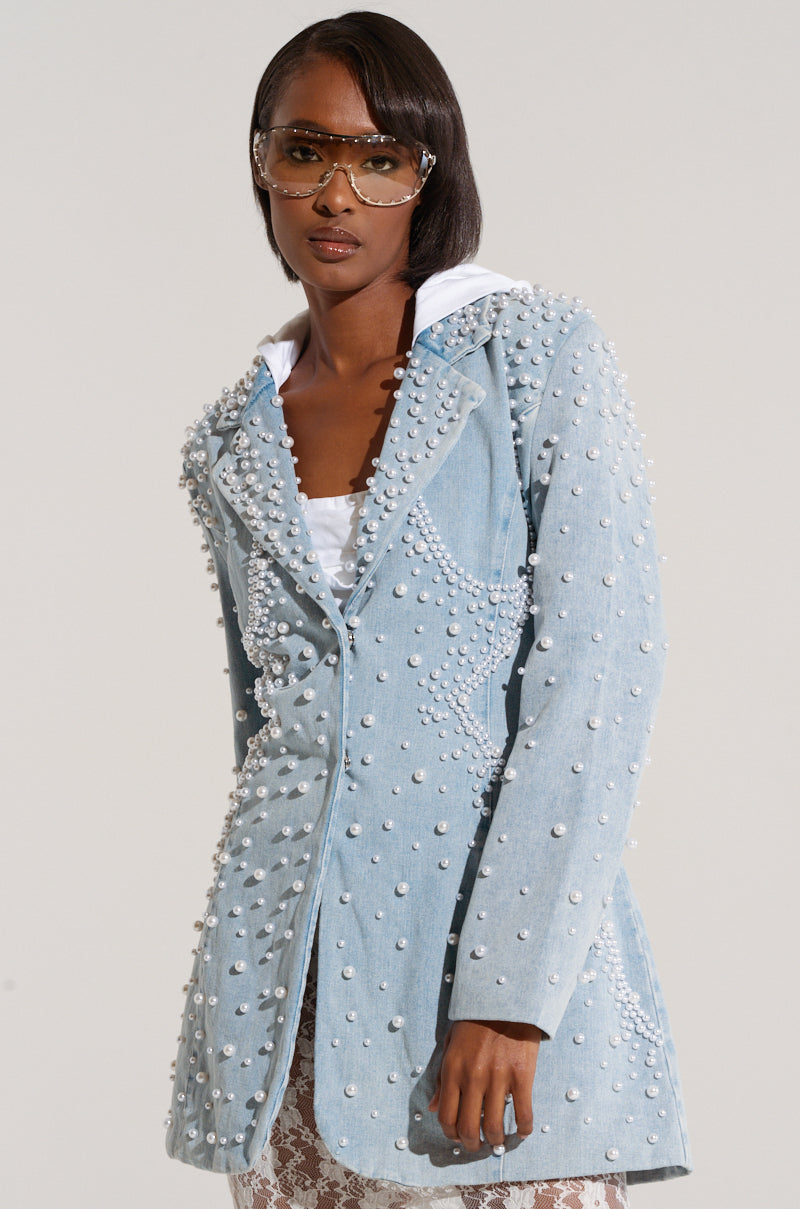A person with short, straight hair is wearing clear-rimmed sunglasses and a light blue ALIX PEARL EMBELLISHED BLAZER from AKIRA Label over a white top. They are posing against a plain background.
