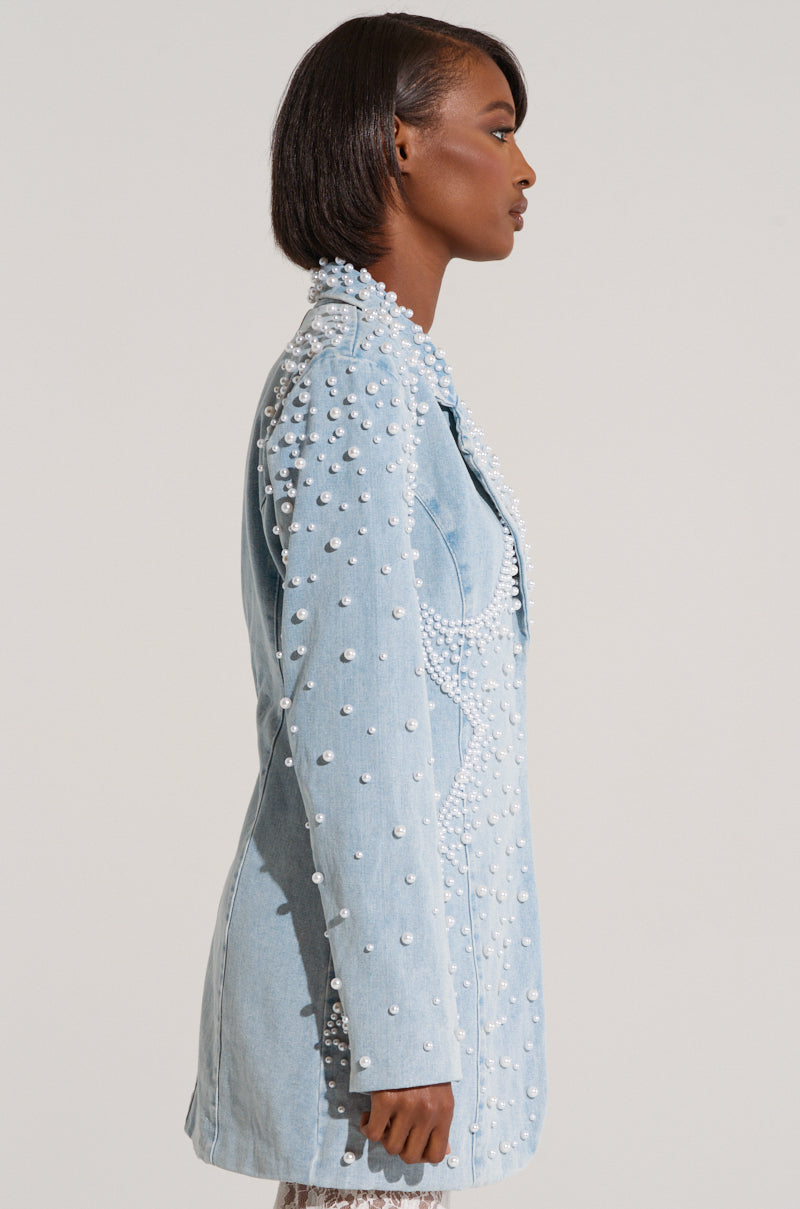 A person with short dark hair stands in profile wearing a light blue ALIX PEARL EMBELLISHED BLAZER from the AKIRA Label collection. The background is plain and light-colored.