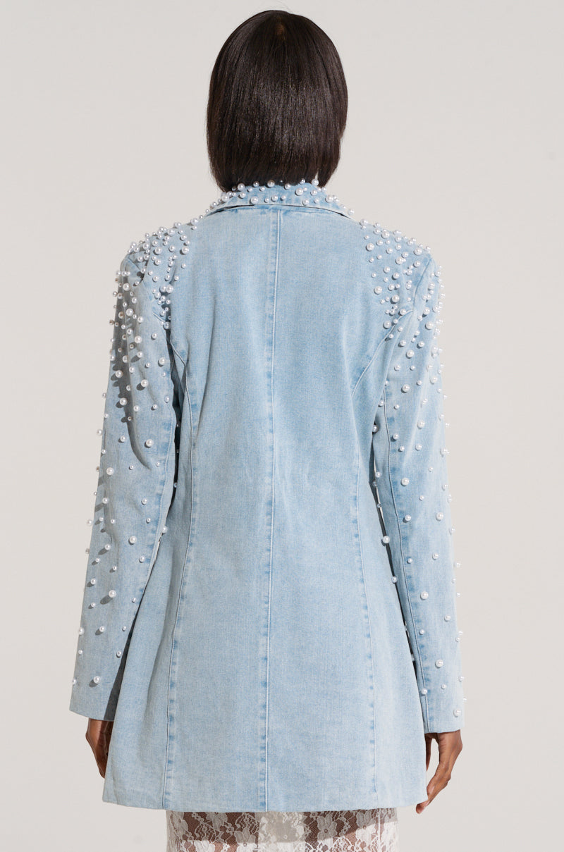 A person with dark hair is shown from the back, wearing an ALIX Pearl Embellished Blazer from the AKIRA Label. This light blue denim blazer is adorned with numerous white pearls along the shoulders and sleeves. The contemporary design of this blazer reaches just below the hips.