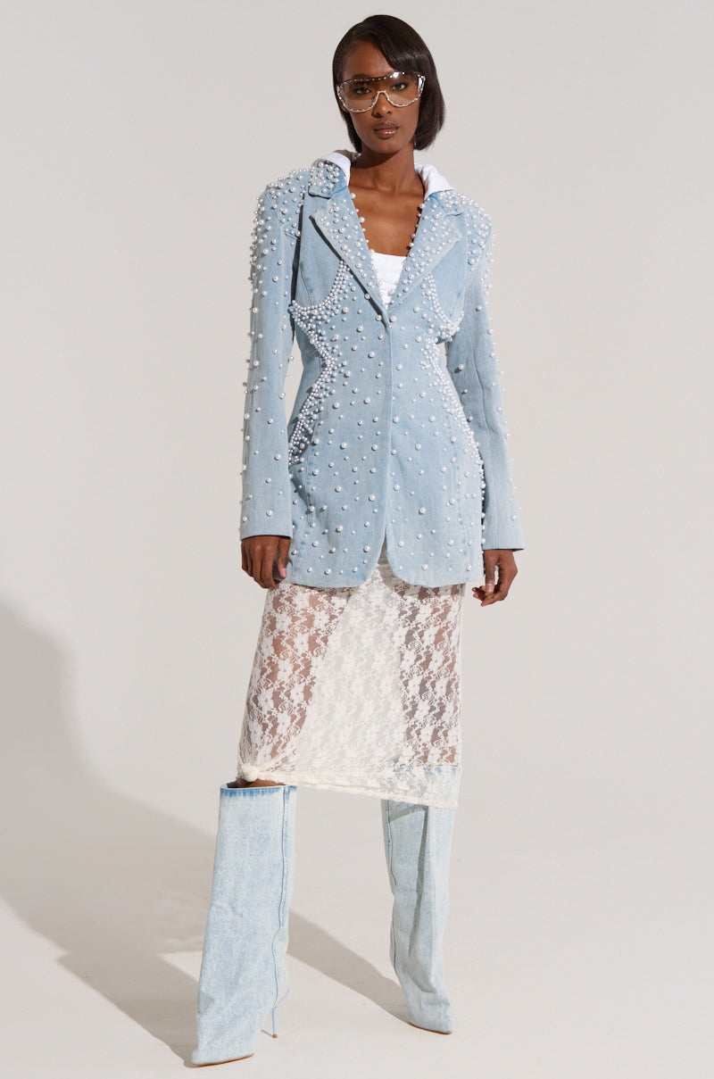 A woman stands confidently in a studio setting, wearing an ALIX Pearl Embellished Blazer over a white outfit composed of a lace dress and matching knee-high denim boots. She completes her look with stylish sunglasses and a sleek hairstyle.