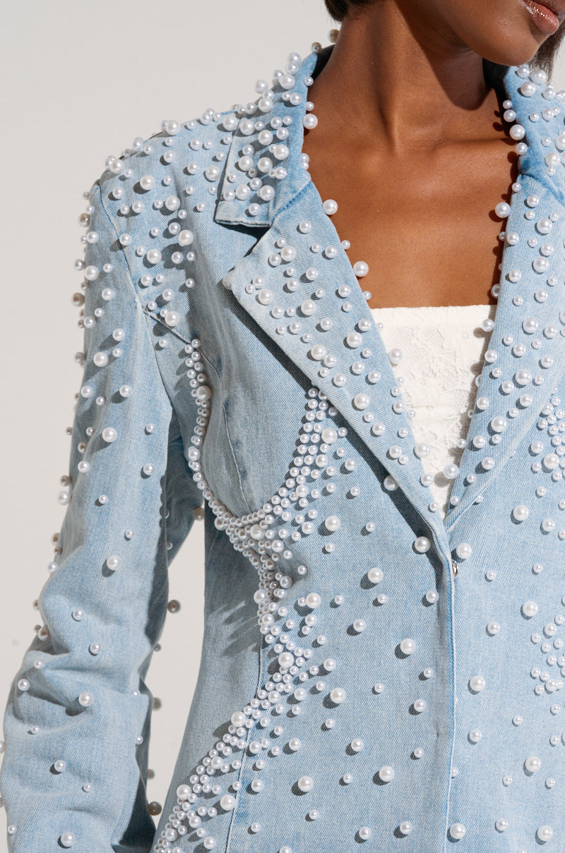A close-up of a person wearing the chic ALIX PEARL EMBELLISHED BLAZER from the AKIRA Label collection. The light blue denim blazer is adorned with numerous white pearls, scattered across the sleeves, lapels, and front to form intricate patterns. Underneath the blazer, the person wears a white strapless top.