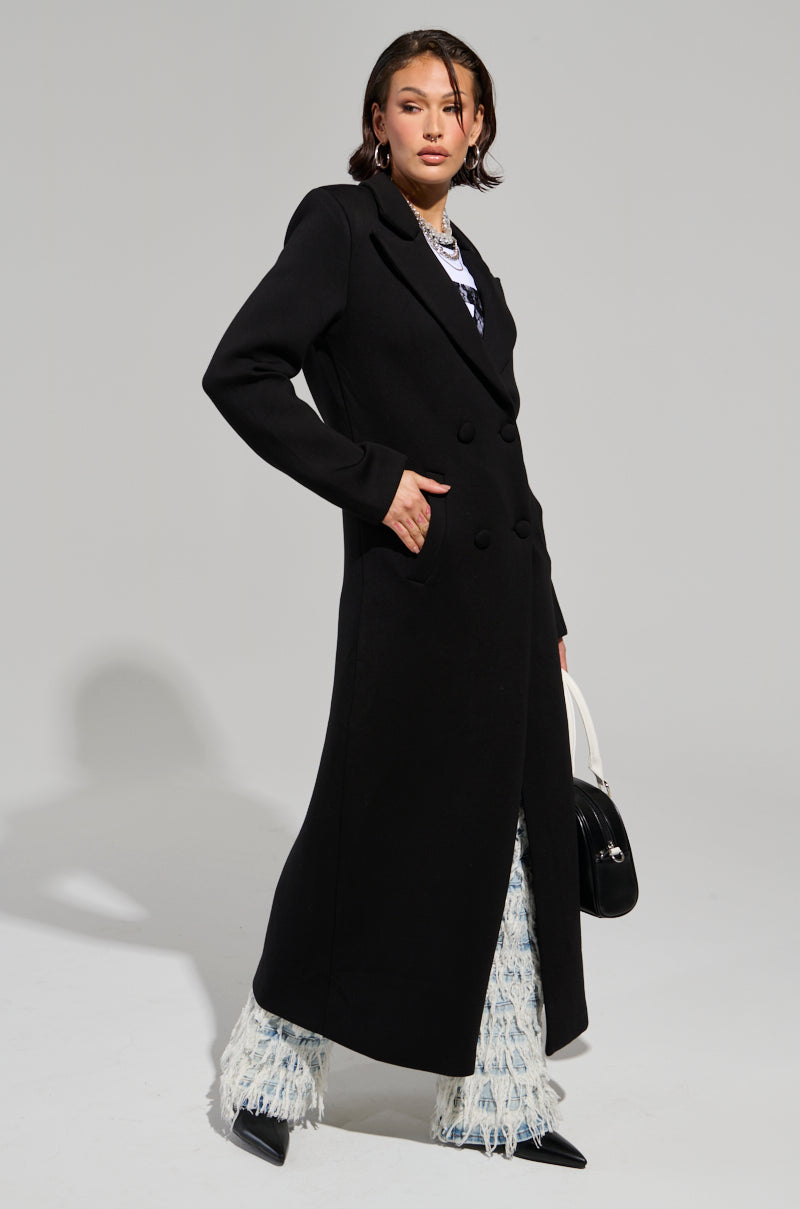 A person poses confidently in a double-breasted overcoat, identified as the COCOA BUTTER SCUBA TRENCH IN BLACK, paired with white patterned pants and black pointed shoes. They complement the look with a matching handbag and hoop earrings. The well-lit background enhances the elegance of the trench.