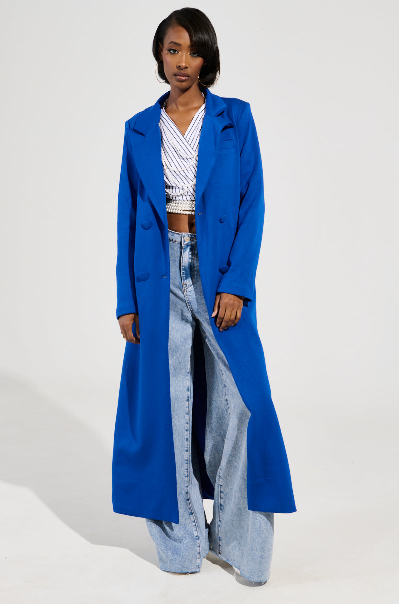 A person wearing a COCOA BUTTER SCUBA TRENCH IN BLUE over a striped crop top and wide-leg jeans stands against a neutral background. They have short dark hair and a confident expression.