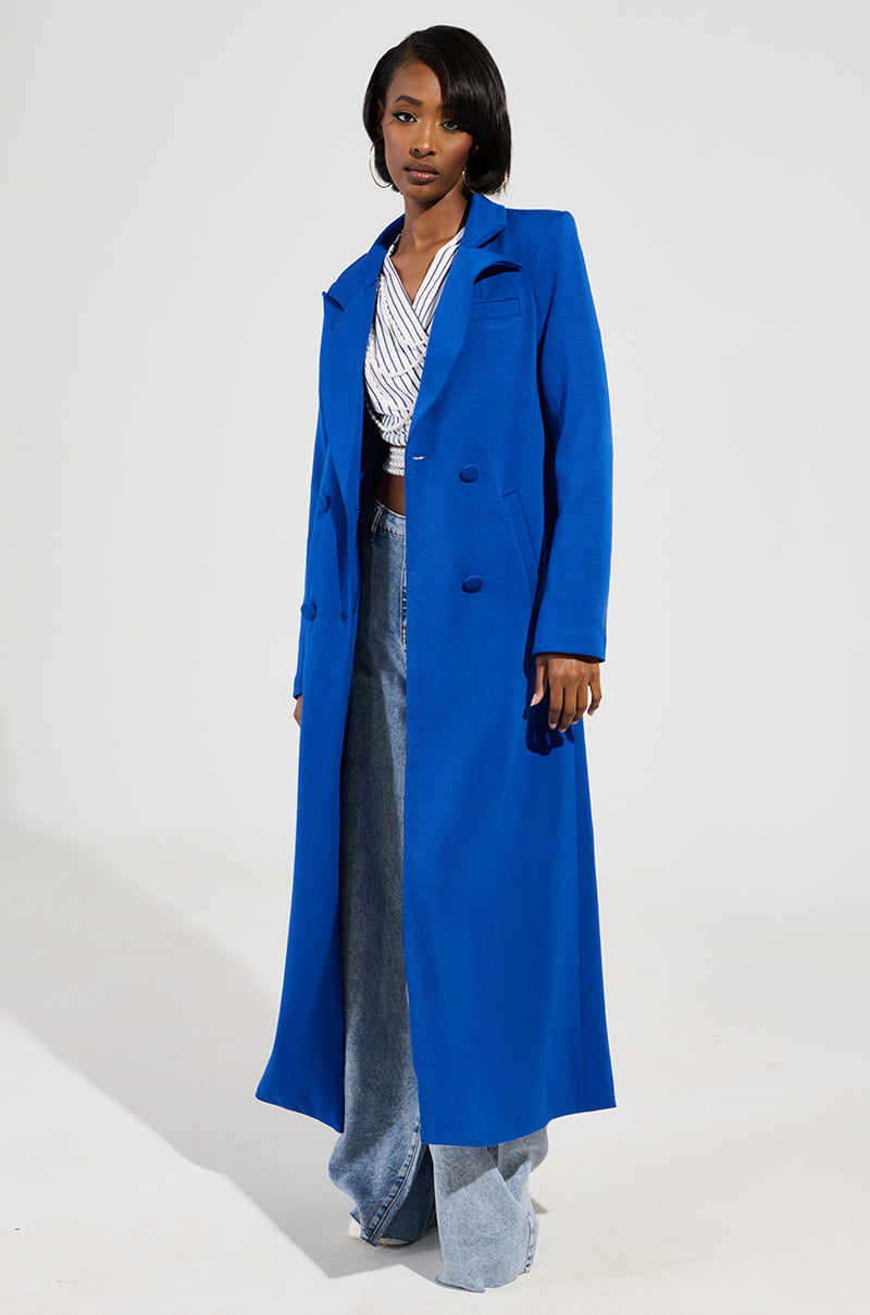 Person wearing a vibrant Cocoa Butter Scuba Trench in blue over a white and blue striped top and wide-leg jeans, standing against a plain background.