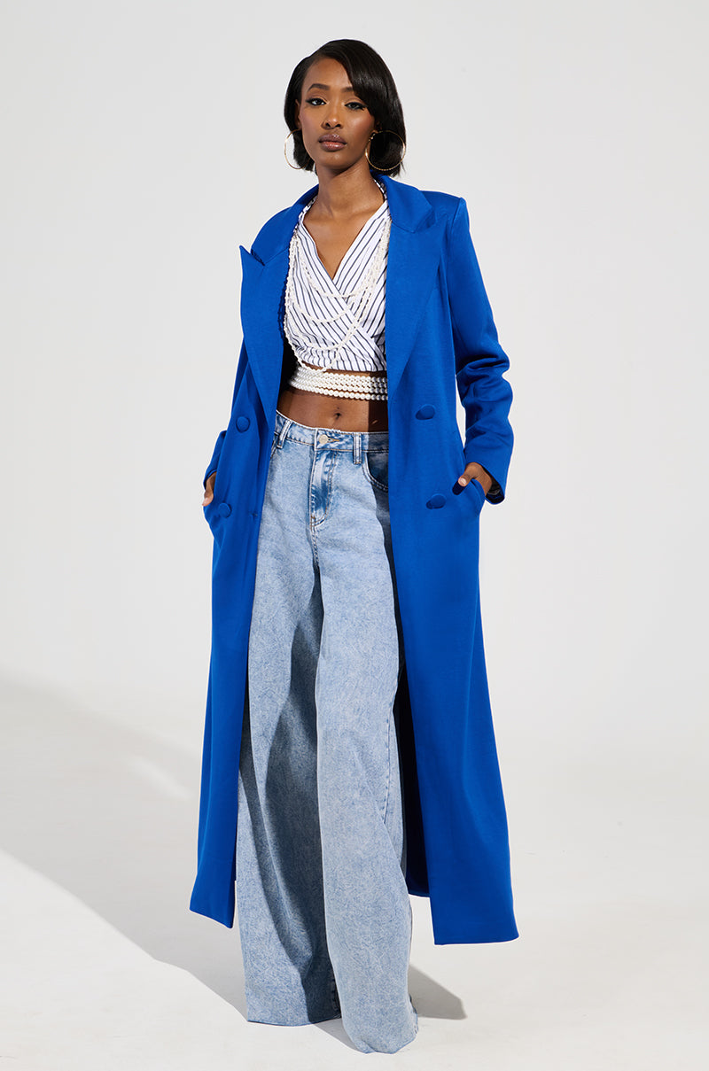 A person wearing a COCOA BUTTER SCUBA TRENCH IN BLUE from AKIRA Label, a white patterned wrap top, and light blue wide-legged jeans stands against a plain background. They have hands in pockets and gaze confidently forward.