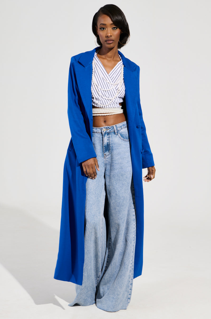 A person wearing a vibrant COCOA BUTTER SCUBA TRENCH IN BLUE over a white textured top and wide-legged light blue jeans stands confidently against a plain white background, showcasing their stylish demeanor.