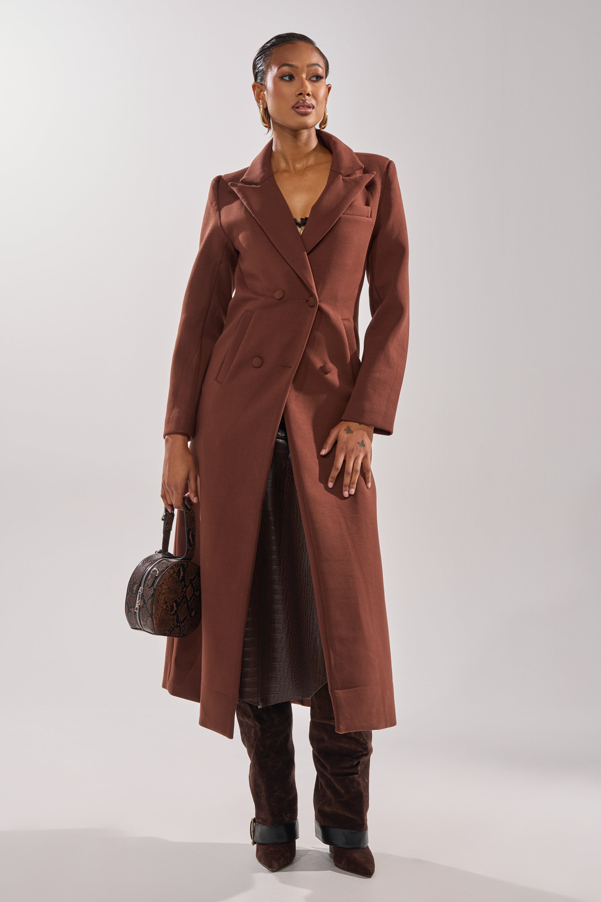 A woman wears the COCOA BUTTER SCUBA TRENCH IN BROWN, paired with brown heeled boots and a textured brown handbag. She accessorizes with hoop earrings, her hair styled back, against a plain, light background. 0