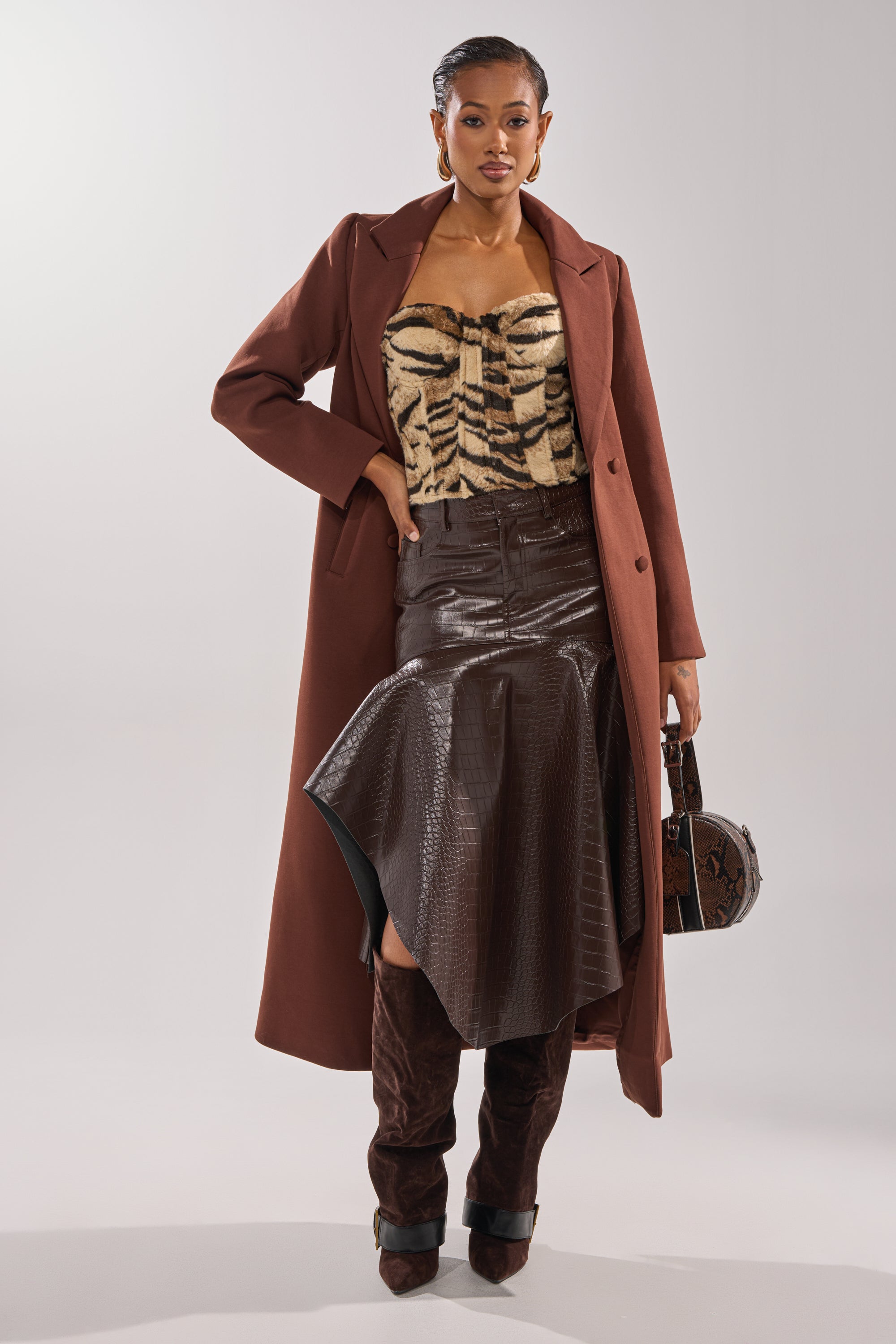 A woman stands facing forward in the COCOA BUTTER SCUBA TRENCH IN BROWN, layered over a tiger-print top, an asymmetrical dark brown leather skirt, and knee-high boots. She holds a round brown handbag with her hair slicked back. 1