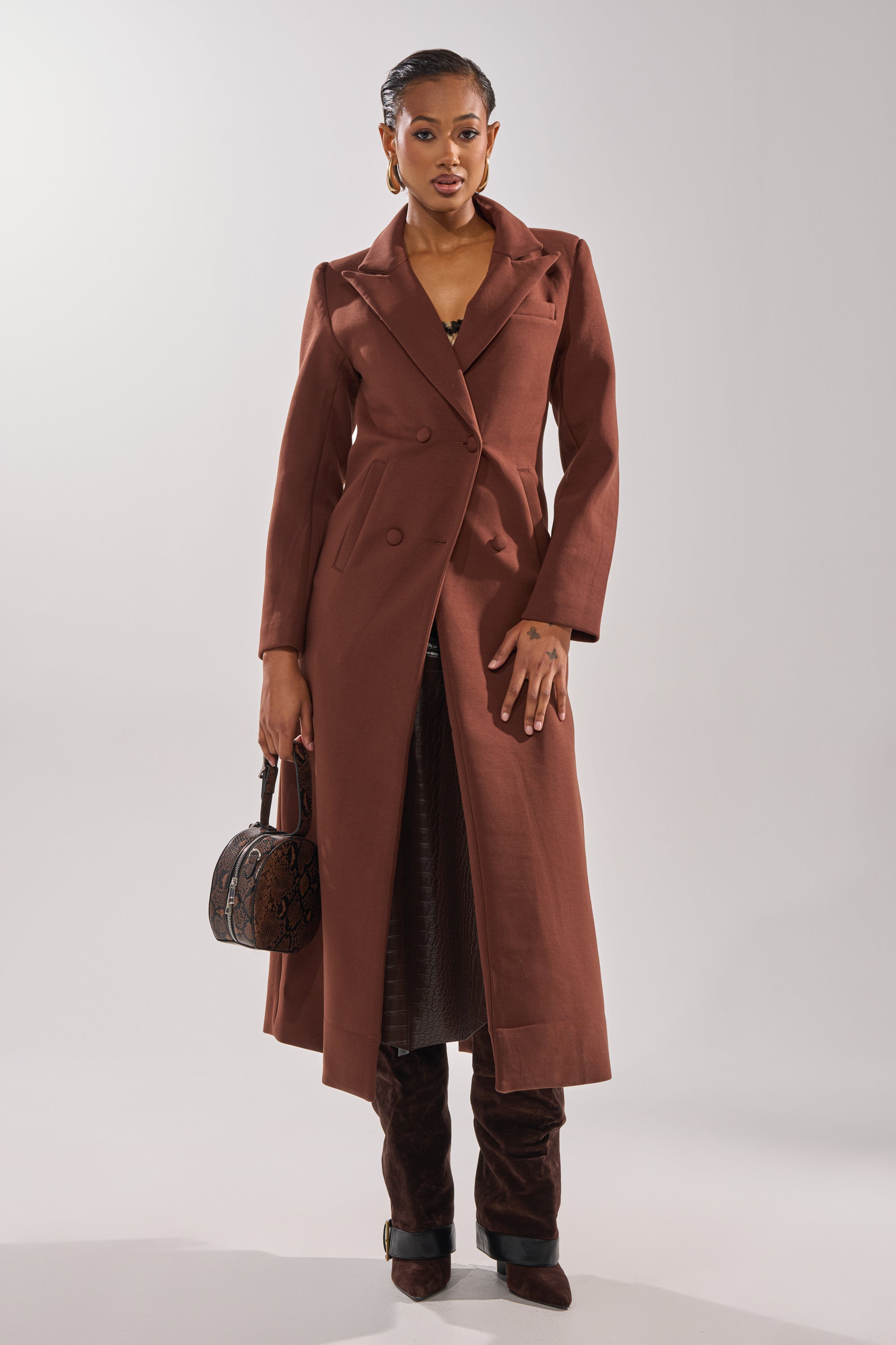 A woman with slicked-back hair and a confident expression stands against a plain background, wearing the COCOA BUTTER SCUBA TRENCH IN BROWN, dark brown boots, and holding a textured brown handbag. 2