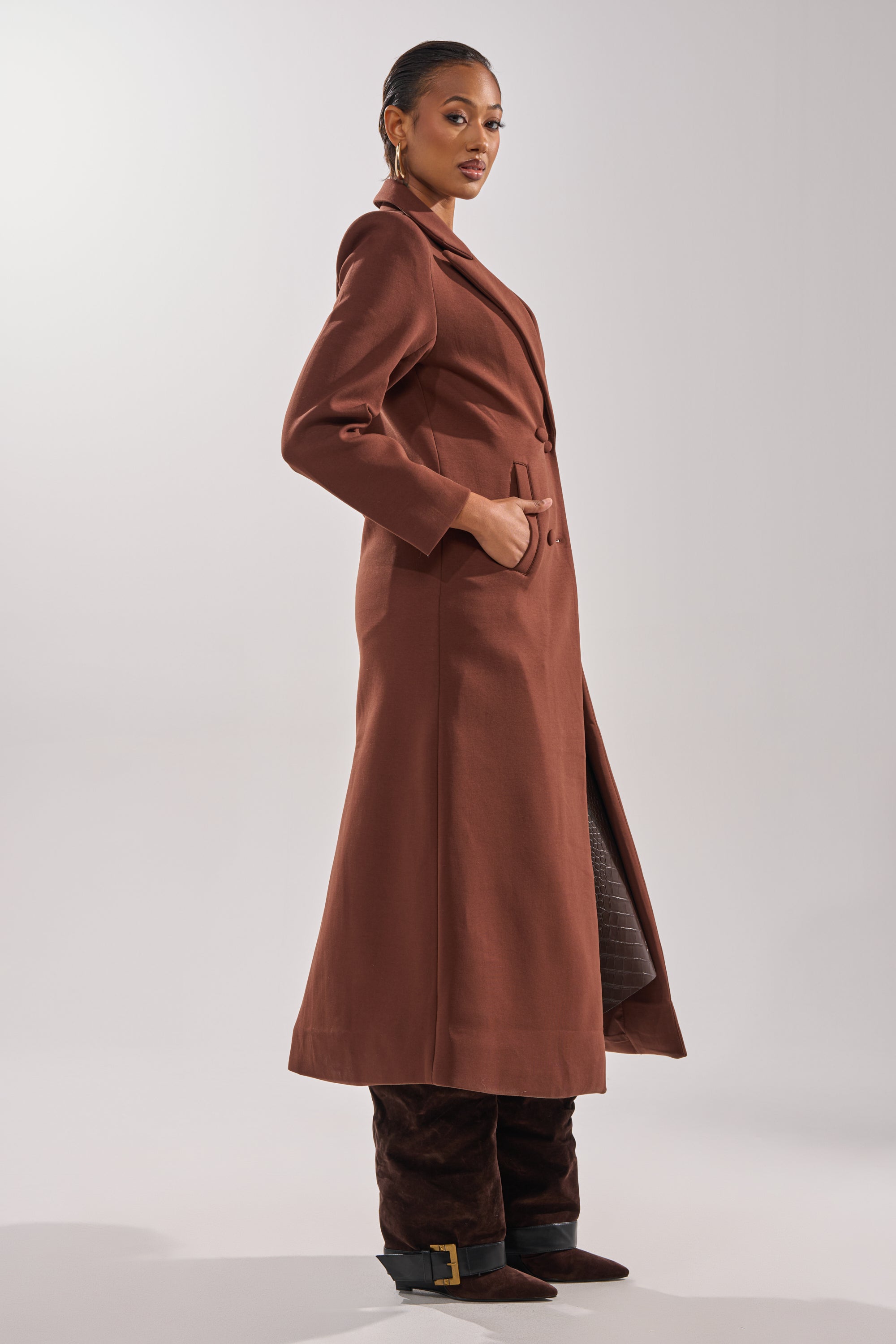 A woman stands in profile wearing the COCOA BUTTER SCUBA TRENCH IN BROWN, her hand in the pocket, styled with dark brown knee-high boots and large hoop earrings against a plain light background. 3